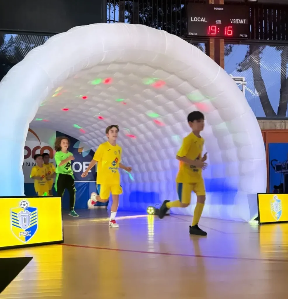 Jóvenes deportistas con equipación amarilla atravesando un túnel de entrada inflable iluminado durante la presentación de un equipo en un pabellón.
