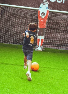 Niño practicando puntería lanzando un balón de fútbol hacia una lona con objetivos de puntuación en un campo de césped artificial.