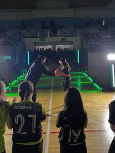 Dos jóvenes deportistas de espaldas frente a una estructura de gradas iluminadas con luces verdes durante una ceremonia de apertura deportiva.