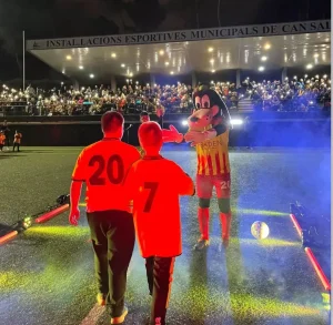 Dos jugadores con equipación roja caminando sobre una pista exterior decorada con efectos de luces de colores y humo para un evento nocturno.