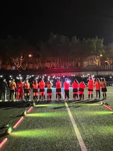 Alineación de un equipo infantil de fútbol con uniformes rojos sobre el césped, iluminados por focos de suelo en una presentación oficial nocturna.