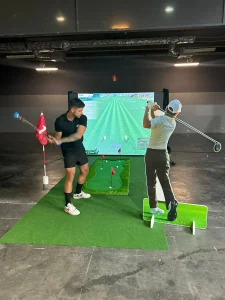 Vista de un simulador de golf en interiores con un jugador en posición de swing y un soporte gráfico que muestra la trayectoria de la bola.